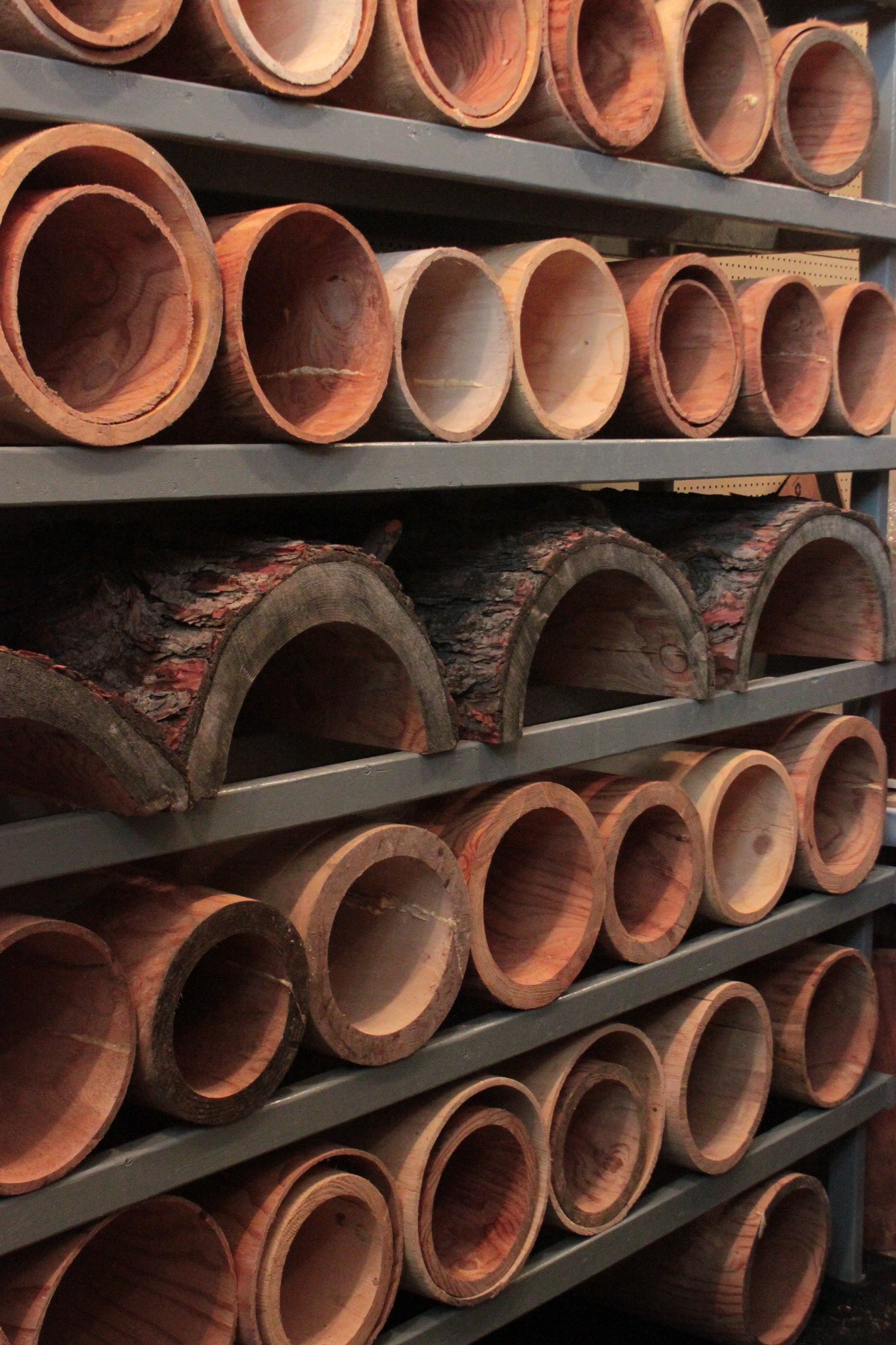 Floor-to-ceiling steel shelving rack packed with finished IC WOOD hollow cylinders and bark-on half-log pieces — warm red pine grain, production warehouse, March 2014